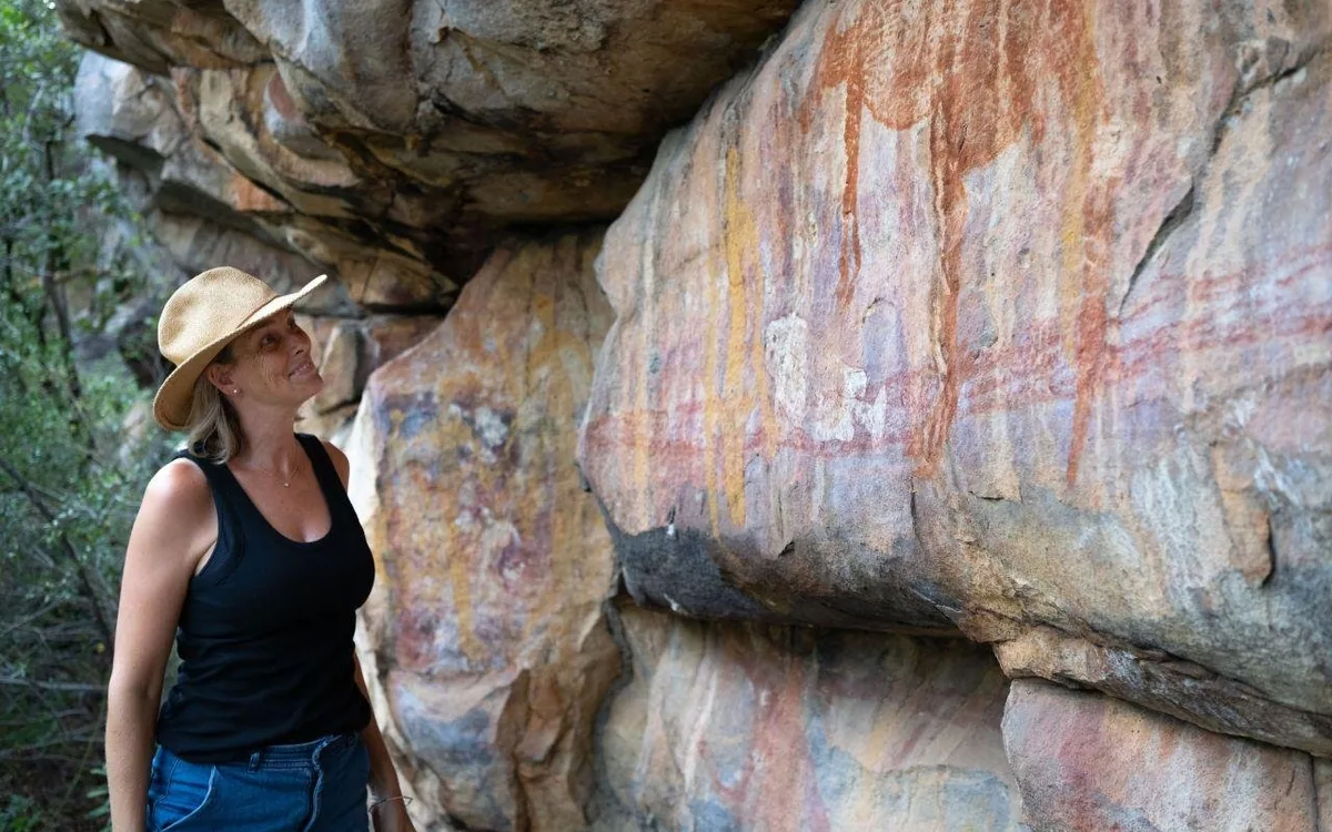 A woman in a hat looking up at ochre figures painted on a rock face under an overhang