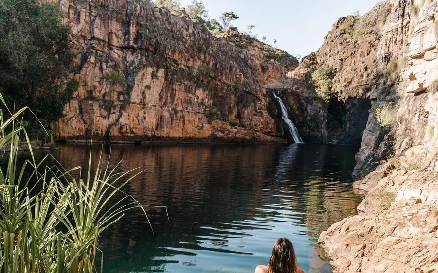 A waterfall dropping into a deep green pool below a red cliff, reeds in the foreground