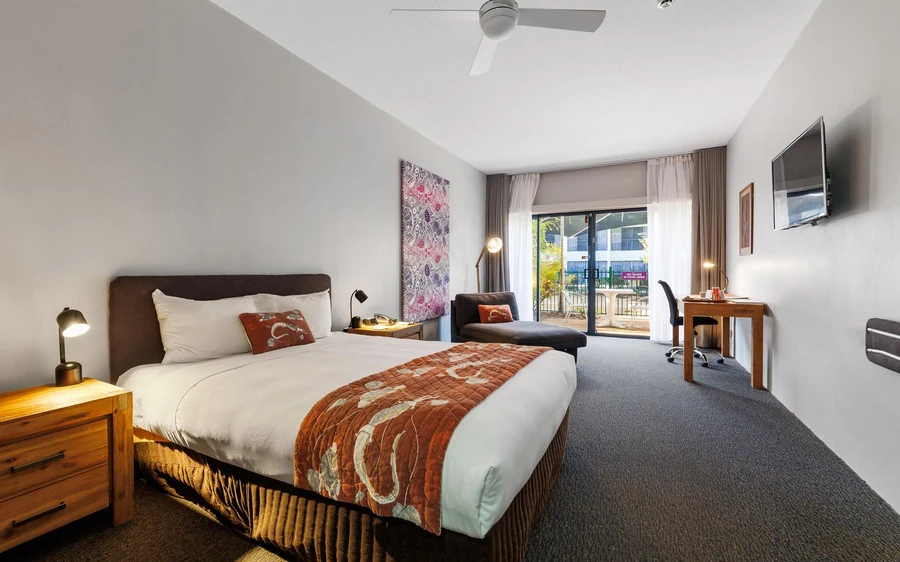 A made-up bed, a desk and a wide balcony door in a plain, bright room