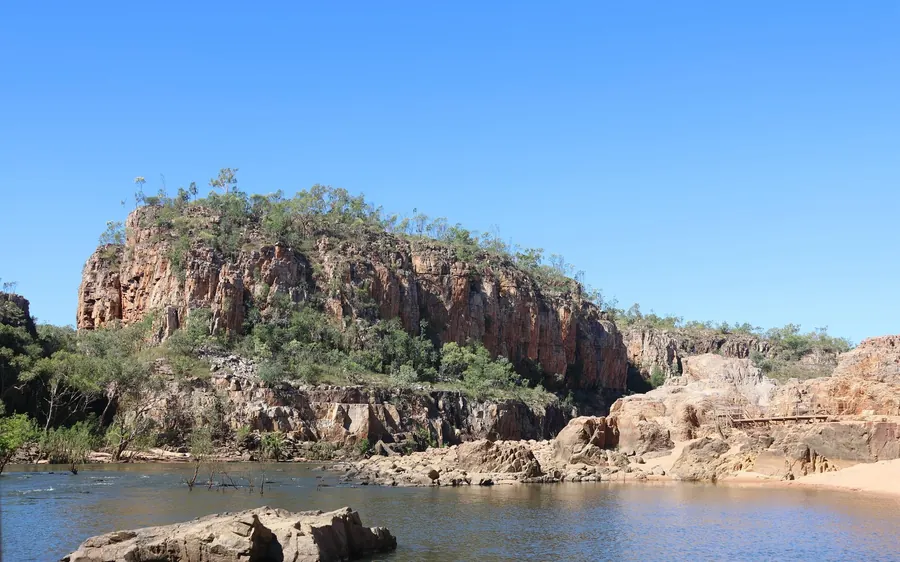 A red sandstone bluff with trees on top above the calm river, a sandbank on the far side