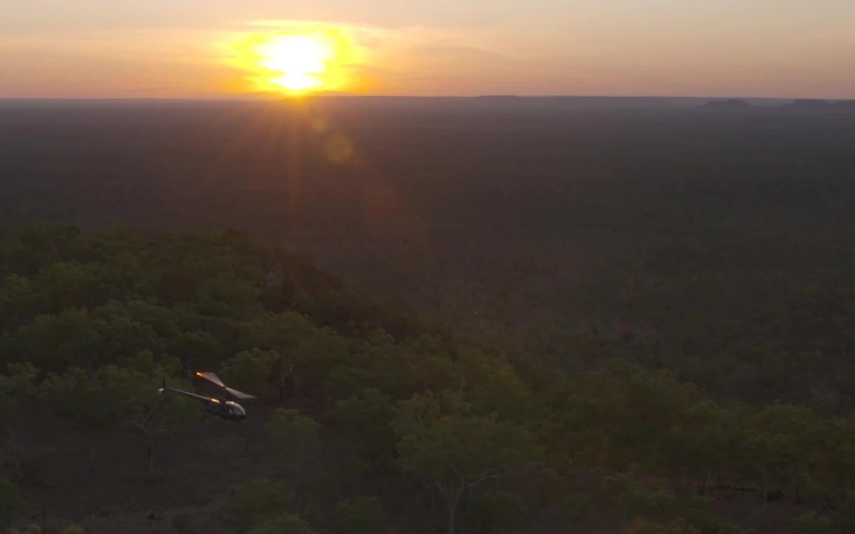 A helicopter low over wooded plateau country as the sun drops to the horizon